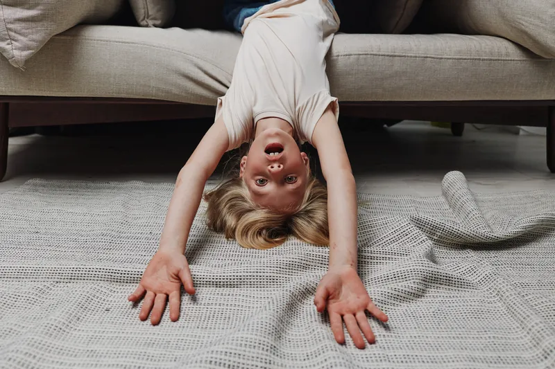 Happy child lying upside down on carpet with arms stretched out under sofa, playful mood in cozy living room concept of childhood fun and relaxation.