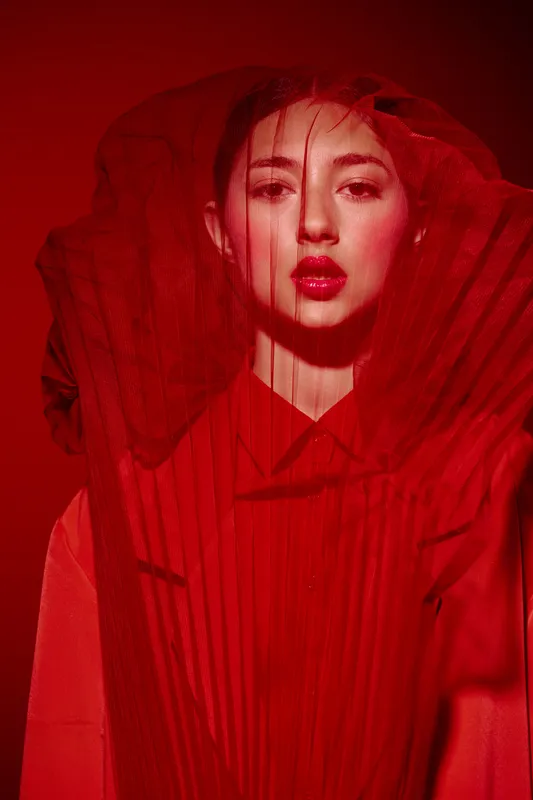 Fashion model in vibrant red outfit and sheer fabric veil posing dramatically. Bold red lips complement the creative and avant-garde beauty concept presented in studio lighting.