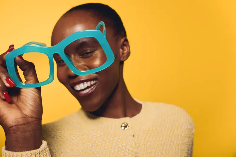 Smiling dark skinned black woman holds large blue glasses frame near face on yellow background. Concept of beauty, fashion, joy, and playful expression with vibrant studio lighting.