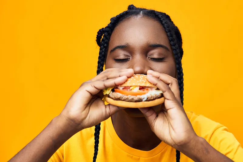 Young woman enjoying a delicious burger against a vibrant yellow background, showcasing joy and excitement in her expression, wearing a casual yellow t shirt