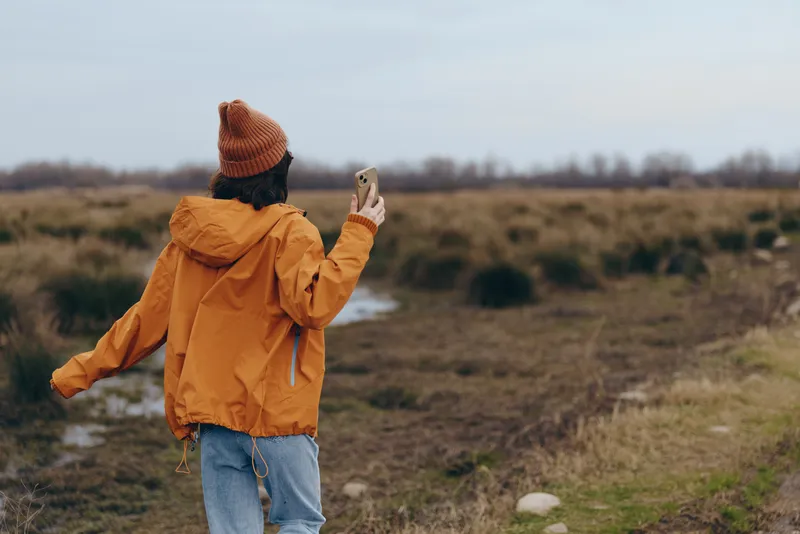 Woman lifestyle outdoors in countryside field, orange jacket hood up, standing on dirt path, holding smartphone for selfie, visible hair, calm mood, distant trees blurred, cloudy sky