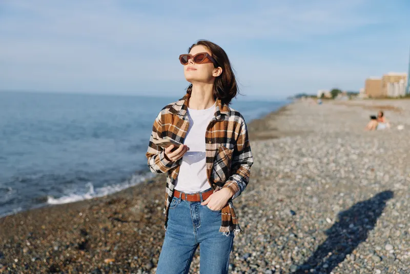 Woman on a pebble beach wearing a plaid shirt over a white tee and jeans, sunglasses, hands in pockets, sunlit sea backdrop, casual fashion portrait by the shore.