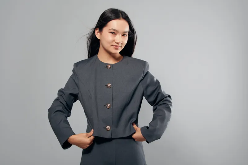 Young woman, portrait and confident pose with hands on waist wearing gray blazer and skirt, studio background. Asian female model with long black hair and casual expression in fashion outfit.
