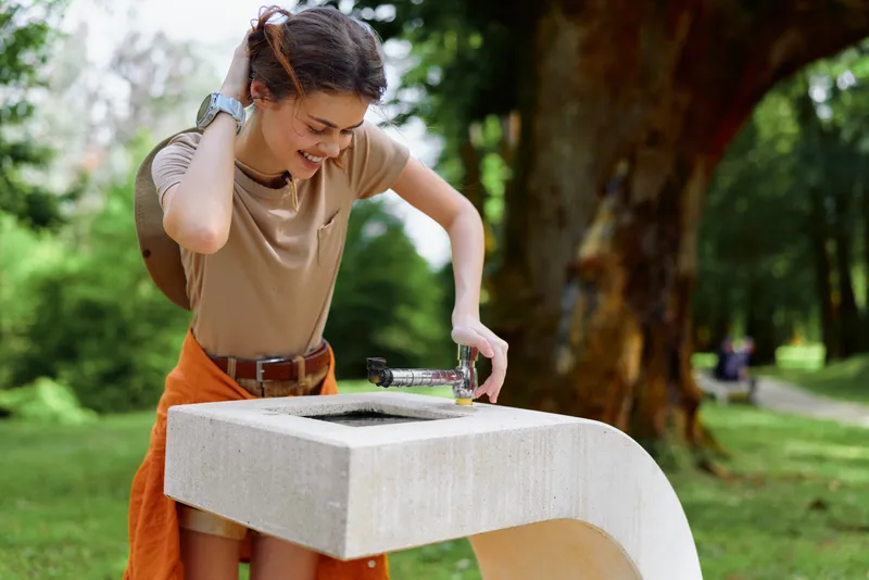 Woman drinking water from a public fountain in a green park, pouring bottle into basin while smiling, casual summer outfit, outdoor leisure and hydration moment.