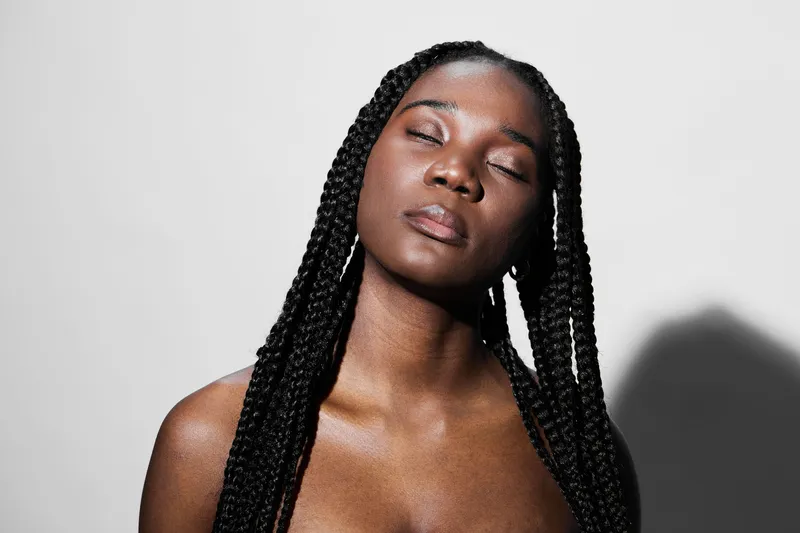 A serene woman with braided hair and closed eyes, expressing calmness and confidence against a minimalist white background, perfect for beauty and self care themes