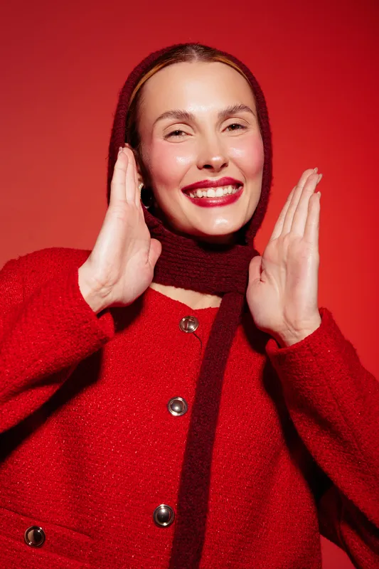 Smiling woman in red sweater and scarf poses with hands near face on red background. Studio portrait with bright lighting and cheerful mood. Concept of fashion, winter, and happiness.