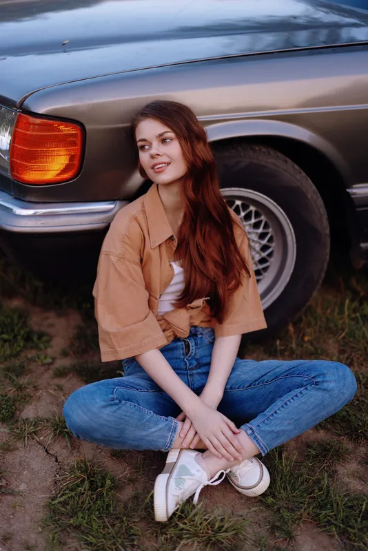 Young woman with long hair in casual attire sitting by vintage car, expressing joy and relaxation in a natural outdoor setting, perfect for lifestyle and travel concepts.
