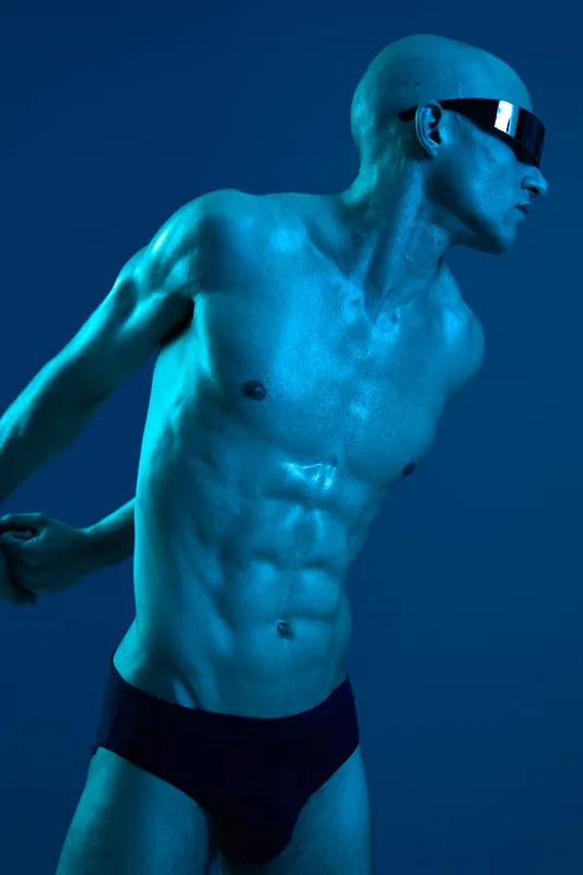 Dynamic man posing with a striking blue light accentuating his muscular physique, wearing sunglasses and minimalist swimwear, showcasing a modern and artistic concept.