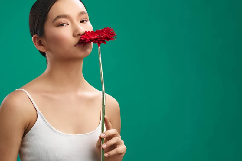Asian woman stands against a vivid backdrop, gently smelling a bright red flower as she poses in a minimalist studio setting, creating a calm, intimate moment with natural beauty and soft lighting.