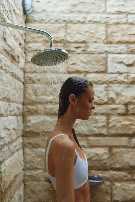 young woman standing under outdoor shower with wet hair, stone wall background, relaxed mood, summer lifestyle, natural light