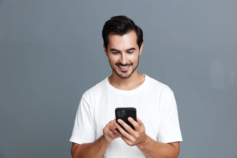 Smiling man in casual white t-shirt looking at smartphone, standing against a plain gray background, conveying joy and engagement with technology.