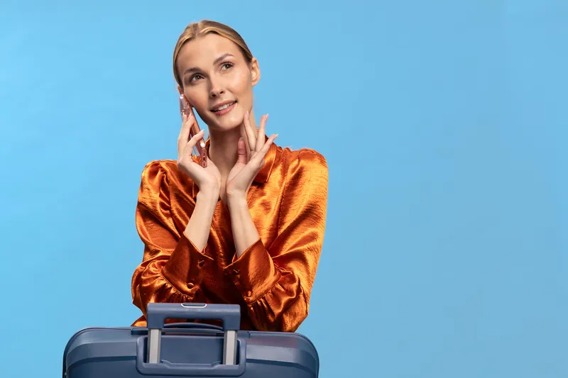 Blonde woman in orange satin blouse poses with blue suitcase on blue background. Concept of travel, tourism, fashion, and lifestyle with elegant female traveler and luggage.