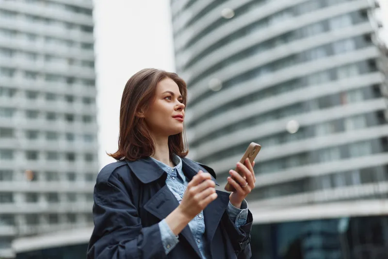 Urban scene shows an adult brunette woman in a navy trench coat looking at her smartphone with a focused smile among glass skyscrapers on a sunny day.