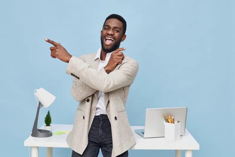 Confident smiling man, young adult African American, casual beige blazer and white shirt, pointing with both hands, standing in modern office with white desk, laptop, and plant, cheerful mood, bright 