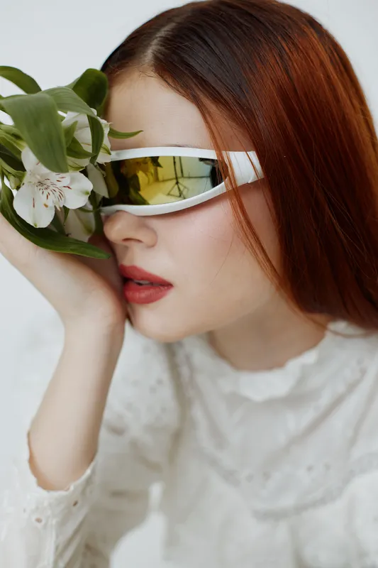 Young woman in futuristic sunglasses with flowers, expressing serene emotions and wearing a stylish white outfit in a soft color palette