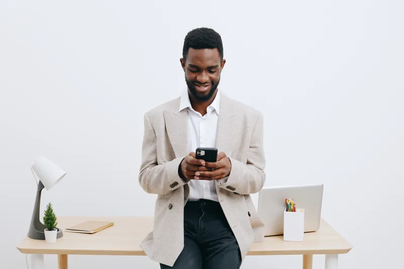 Smiling man using mobile phone in modern office, casual business attire, African descent, positive mood, standing near desk with laptop and stationery, clean minimalistic background. People lifestyle 