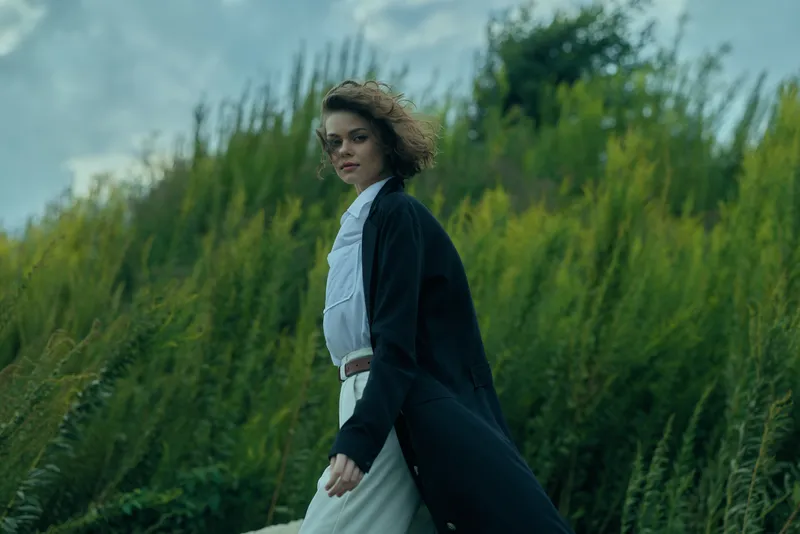 Woman in a long coat walks through tall grasses on a windy day, fashion-forward style and dynamic movement captured in natural light.