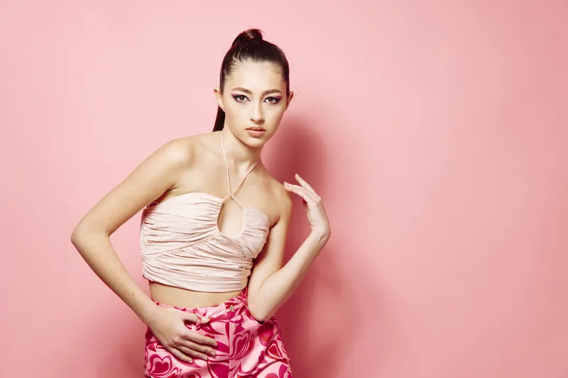 Stylish young woman with dramatic makeup wearing a creative cropped top posing against a pink backdrop. Concept for beauty, fashion, and creative editorial style.