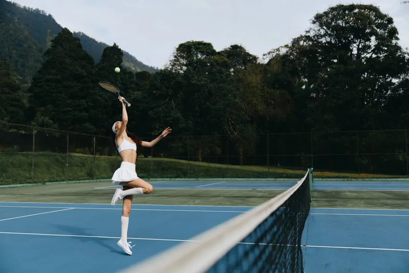 Athletic woman in a white tennis outfit serves on a blue court in a lush green setting, showcasing determination and focus during a sunny day