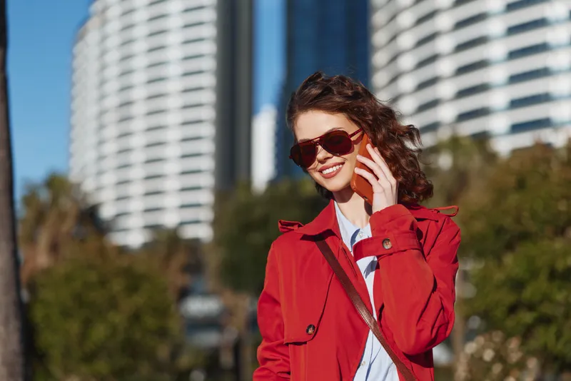 Young woman outdoors wearing red coat and sunglasses talking on smartphone smiling sunny day urban background modern buildings clear sky
