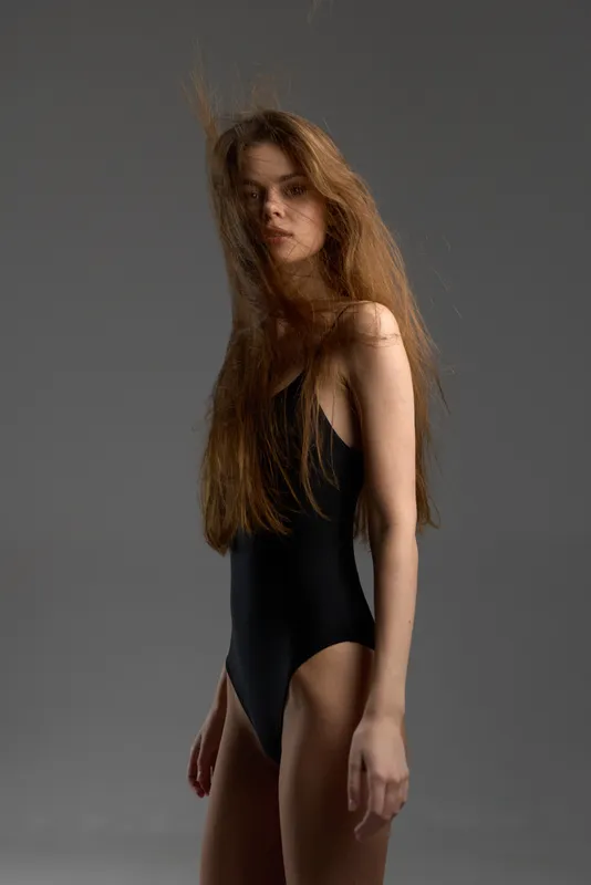 young woman in black bodysuit posing with serious expression on gray background
