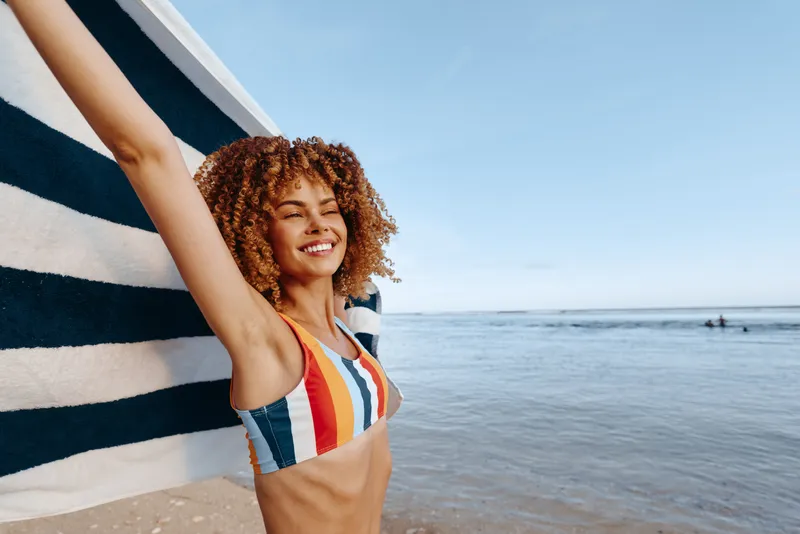 A smiling young woman with curly hair raises her arms beside a striped wall on the beach, capturing a carefree summer moment with the ocean in the background.