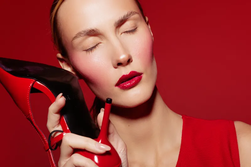 Close-up of a woman with red lipstick and closed eyes holding a red high heel shoe near her face. Red background and dress create a bold, stylish, and elegant fashion concept.