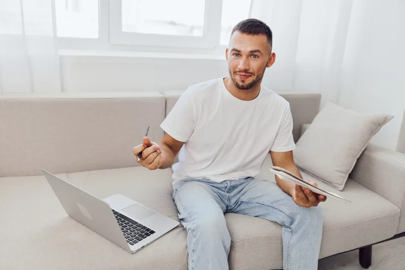 Engaging man sitting on a couch, working on a laptop with a tablet in hand. Bright, modern interior with a cozy atmosphere. Perfect for remote work concepts.