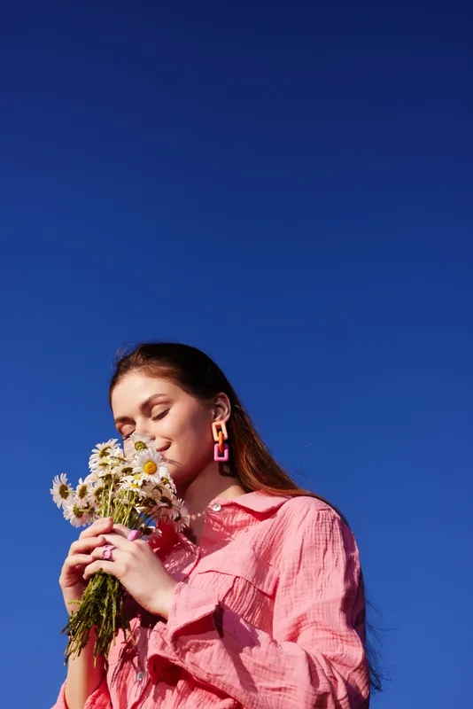 Young woman enjoying a sunny day, holding a bouquet of daisies, wearing a pink dress, smiling against a clear blue sky