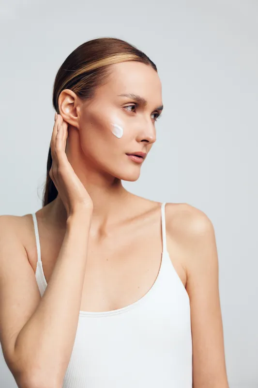 Young woman with smooth skin applying white face cream on cheek, wearing white tank top. Minimalist background emphasizes skincare, beauty, and natural wellness concept with soft lighting.