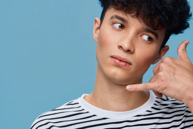 Young man making phone gesture with hand close to face wearing striped shirt against blue background, expressing casual communication and thoughtful mood.