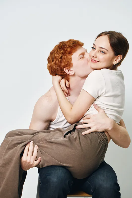 Young couple in love with man holding woman in arms and kissing her cheek. Affection, romance, happiness, close relationship, casual clothes and light background indoors.