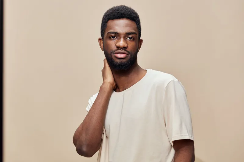 Confident young black man standing against a beige background, wearing a stylish neutral colored t shirt, expressing calm emotions and modern aesthetic