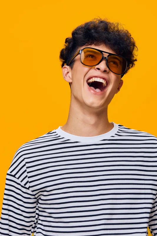 Laughing man wearing striped shirt and large sunglasses against bright orange background, expressing joy and carefree attitude with curly hair and casual style.