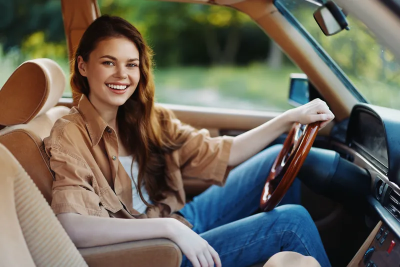 Young woman smiling and driving in a vintage car, showcasing her style and joy in a casual outdoor setting with warm tones and natural light.