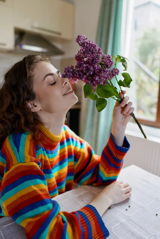 Young woman in a colorful striped sweater leans on a table, smelling a lilac bouquet by a sunlit window. Home lifestyle scene with cozy mood and selfcare moment.