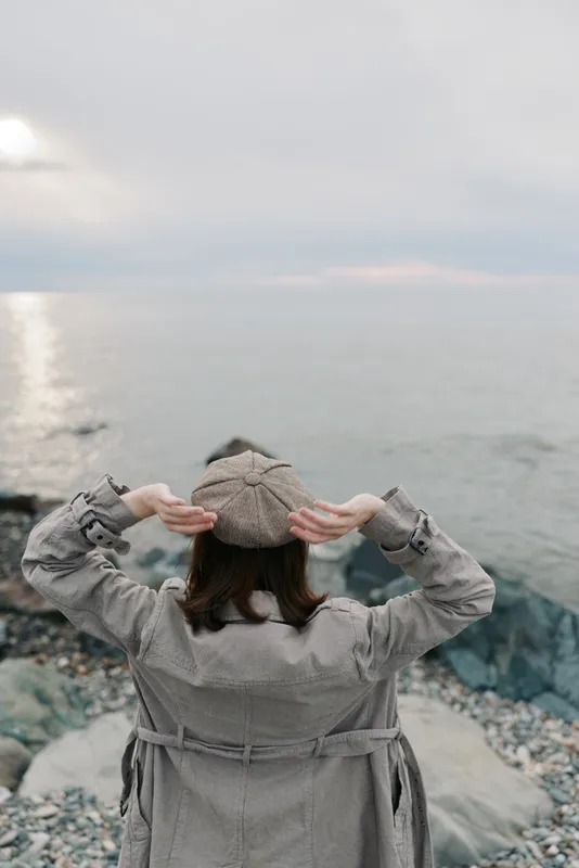 A woman in a grey coat and hat stands on rocky shore facing calm sea under cloudy sky. Concept of solitude, contemplation, and peaceful nature moment by the water.