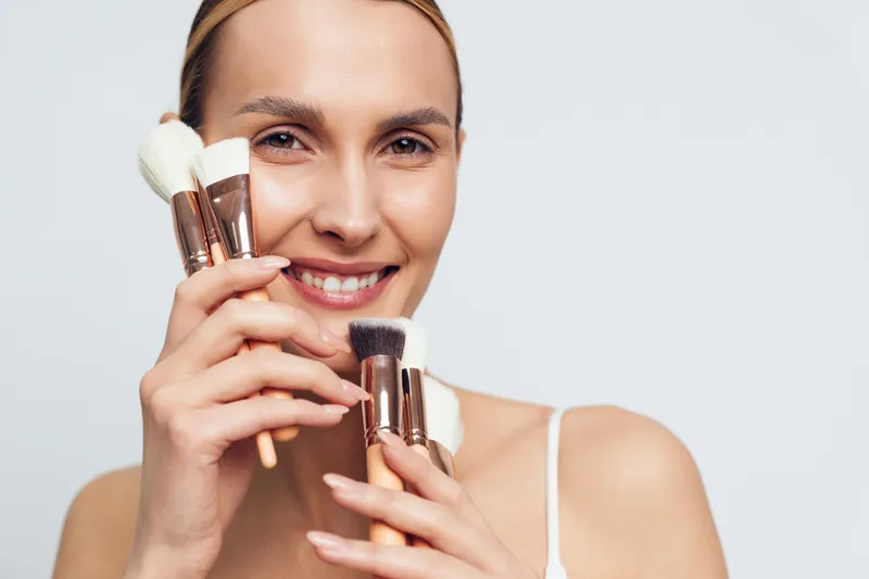 Young woman applying makeup with cosmetic brushes, smiling for beauty close up portrait, holding two foundation brushes near face, clean white background, fresh grooming daily mood.