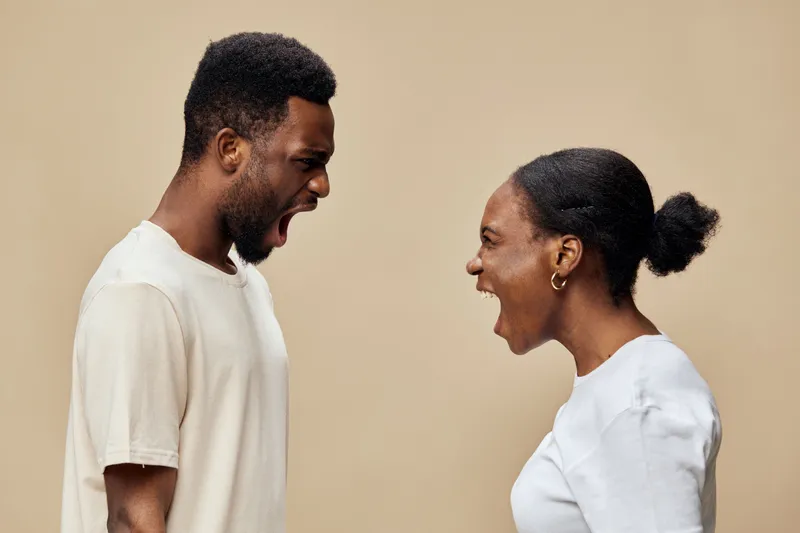 Two young black adults expressing strong emotions while shouting at each other, dressed casually in neutral colors against a beige background, conveying intensity and conflict