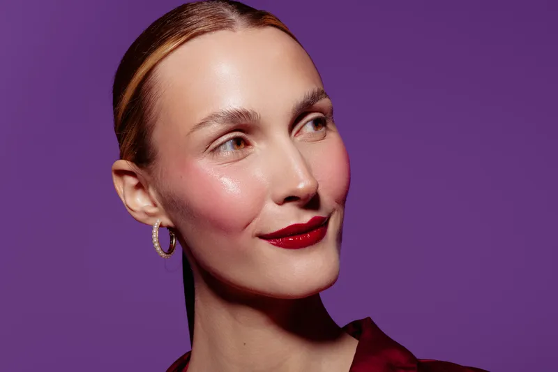 Close-up of a smiling woman with glossy skin and red lipstick looking sideways on purple background. Concept of beauty, makeup, skincare, and confident female portrait.