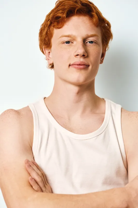 Young man with red hair and freckles wearing white tank top standing with arms crossed on white background. Confident expression, casual style, portrait of male adult with fair skin.