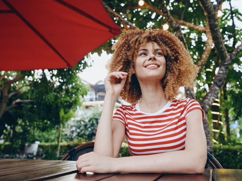 Portrait of a cheerful young woman with curly hair wearing a red and white striped shirt, seated at an outdoor cafe under a bright umbrella, enjoying sunshine.