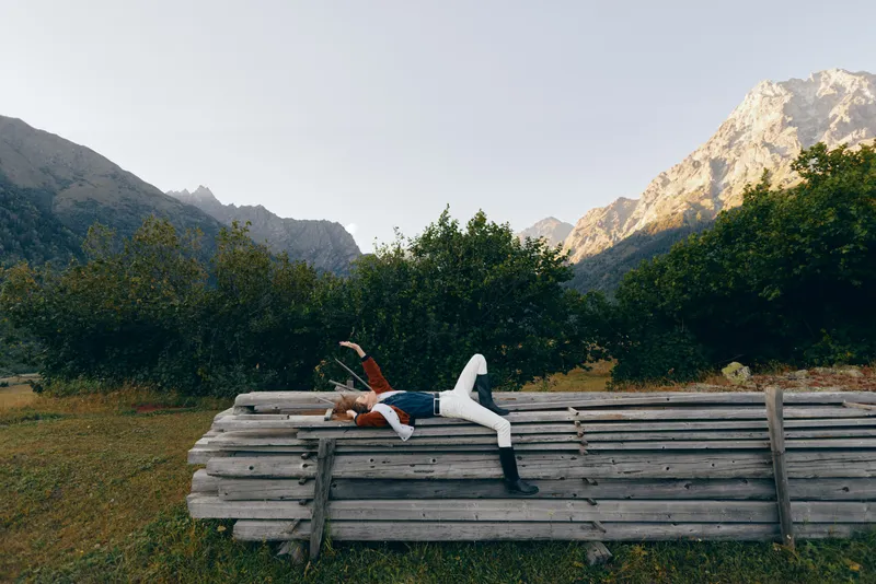 Person woman lying on stacked logs in a meadow, enjoying nature and mountains view, relax outdoors among trees and wooden pile for peaceful countryside rest and leisure.