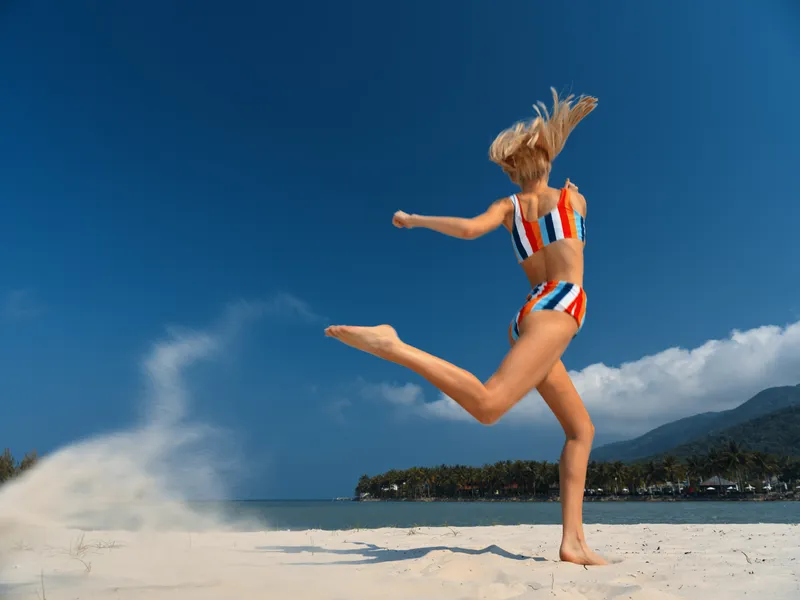 Dynamic action captures a woman mid leap on a sunny beach, wearing a striped swimsuit. Water spray and wind freeze the moment into a bold summer scene.