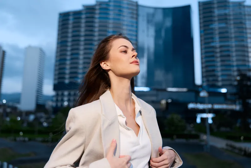 Confident businesswoman in a sleek outfit posing outdoors with modern skyscrapers in the background, exuding ambition and determination.