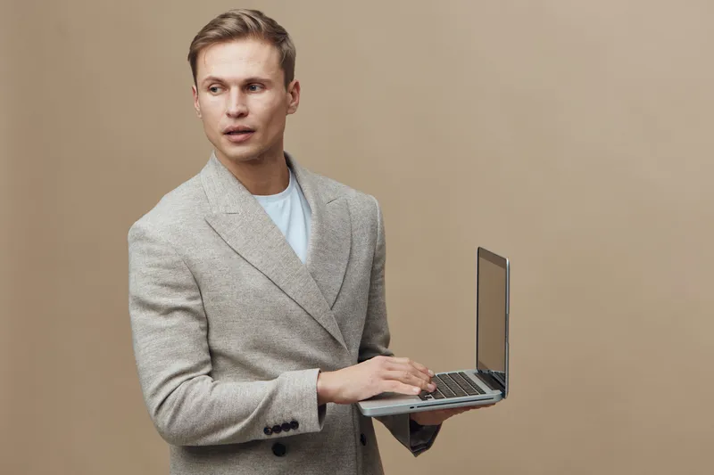 Professional young man in beige suit holding open laptop in hand looking away isolated on plain beige background studio portrait. Business professional concept