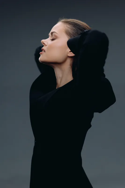 Profile of a calm woman in black clothing with closed eyes and hands behind her head against a dark background.