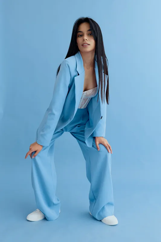 Young woman wearing a light blue suit poses confidently against a matching blue background, showcasing modern fashion and casual style with long straight hair.