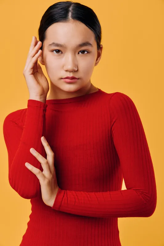 Young woman with black hair wearing red long sleeve top posing with hand near face on orange background. Studio portrait with serious expression and minimal makeup for fashion or beauty concept.