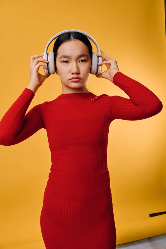 Woman with headphones wearing a red long sleeve dress standing against yellow background for music listening, fashion, and lifestyle. Young adult with serious expression and straight black hair.